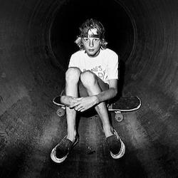 Tony Hawk, as a very, very young man, sitting on his skateboard inside a pipe wearing a snapback and beaten up vans.